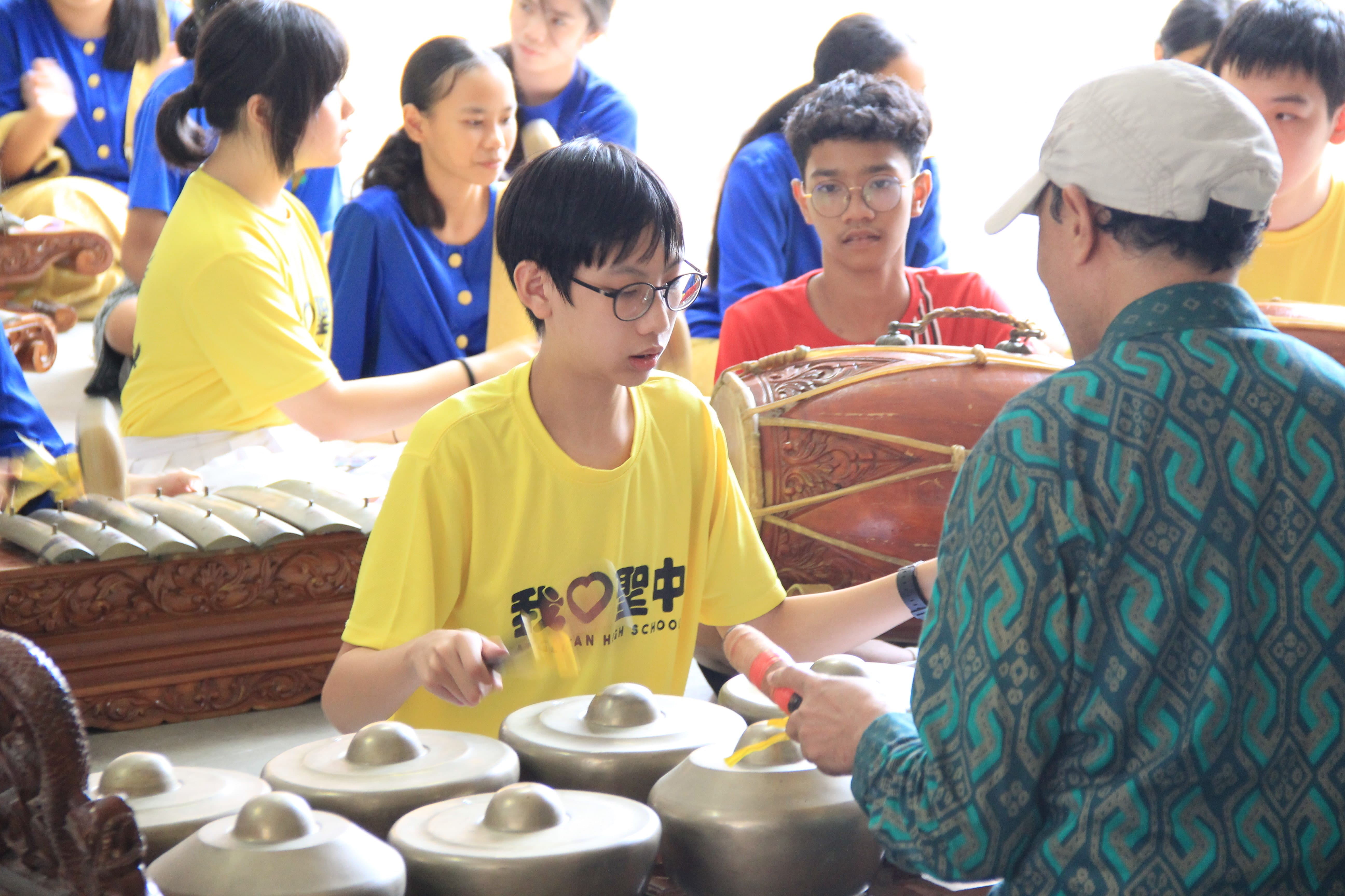 Students in yellow and blue shirts learn to play gamelan instruments guided by an instructor in a patterned shirt.